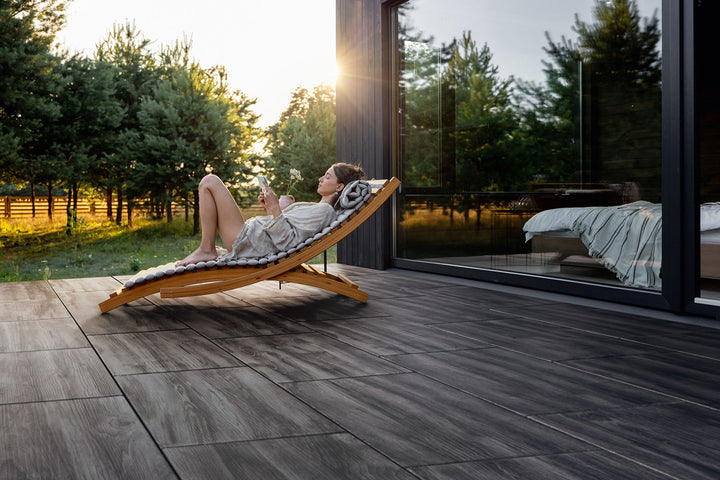 80x40 CreativeLine-Terrassenplatten in grauer Holzoptik von Woehe & Heydemann verlegt auf einer modernen Terrasse mit Person auf einem Liegestuhl