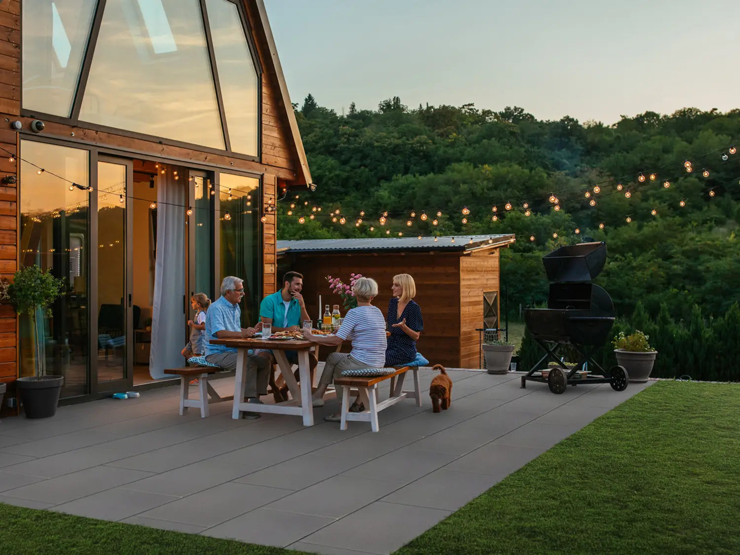 80x40 LuxoGran-Terrassenplatten in grauer Natursteinoptik von Woehe & Heydemann verlegt auf einer gemütlichen Terrasse mit Personen am Esstisch mit Grill und Lichterkette