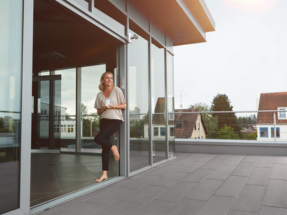 80x40 PremiumLine-Terrassenplatten in grauer Natursteinoptik von Woehe & Heydemann verlegt auf einer Dachterrasse mit Glasfront Frau mit Kaffee