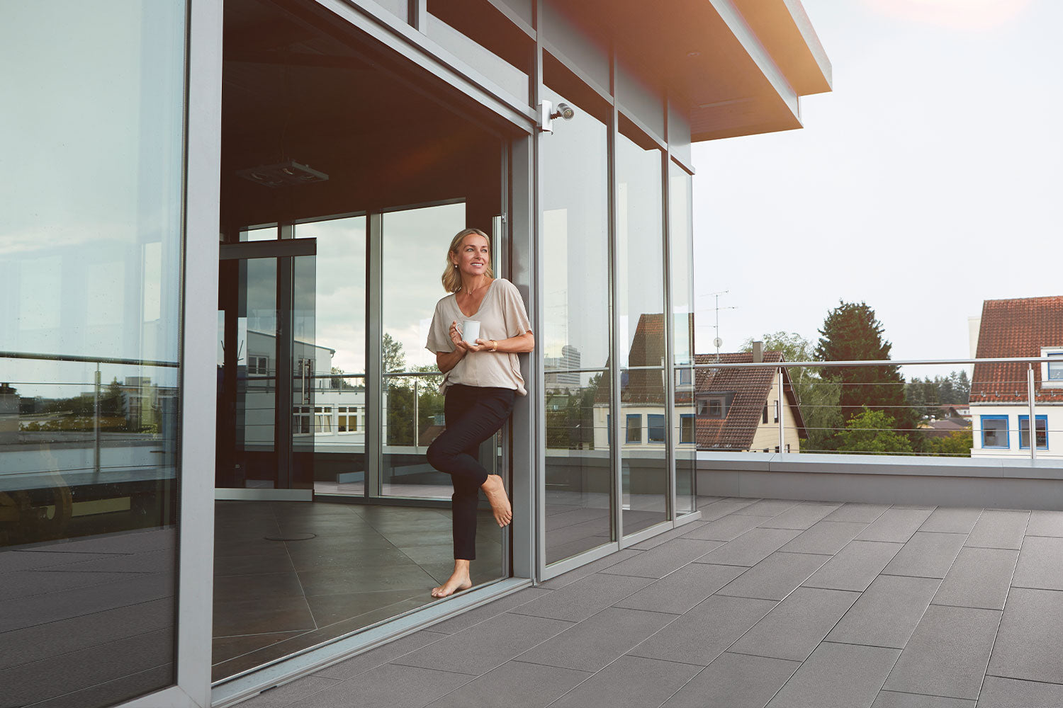 80x40 PremiumLine-Terrassenplatten in grauer Natursteinoptik von Woehe & Heydemann verlegt auf einer Dachterrasse mit Glasfront Frau mit Kaffee